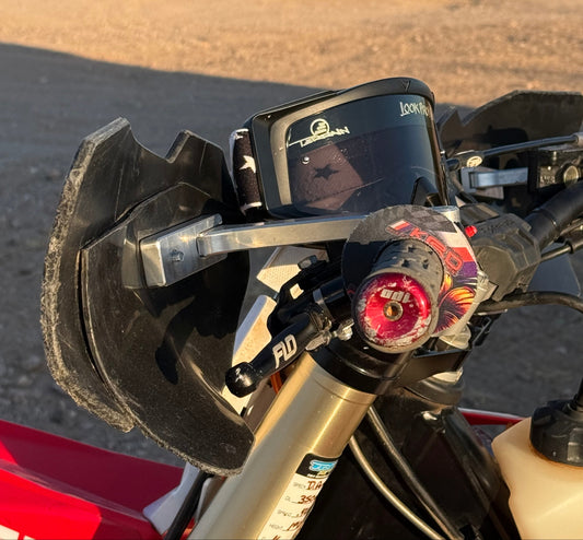 Close-up outdoor shot of a motorcycle handlebar area with a mounted black GPS tracker device on a clear stand, showing part of a red motorcycle body and desert road background.