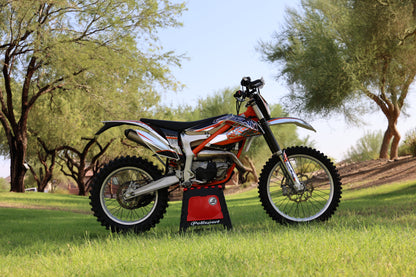A red and black KTM dirt bike is shown on a grassy lawn with trees in the background, supported by a red and black stand branded with 'KTM'.
