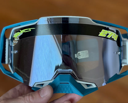 A close-up of motocross goggles held in a hand, featuring a teal frame, a black horizontal stripe across the top, and a reflective silver-tinted lens with lime green branding.
