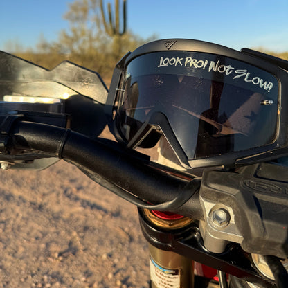 Black motocross goggles with a clear visor featuring the white text 'LOOK PRO! NOT SLOW!' on a desert motorcycle handlebar, with a saguaro cactus and blue sky in the background.
